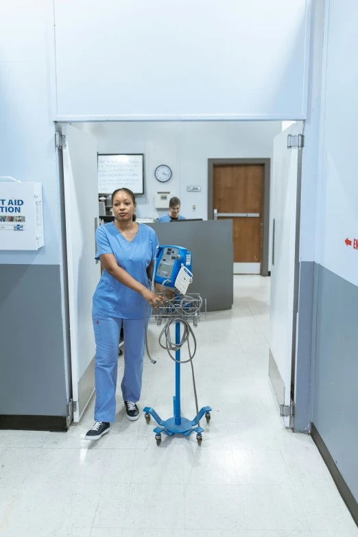 a woman standing next to a machine in a hospital, blue uniform, walking towards the camera, j. h. williams iii, fan favorite