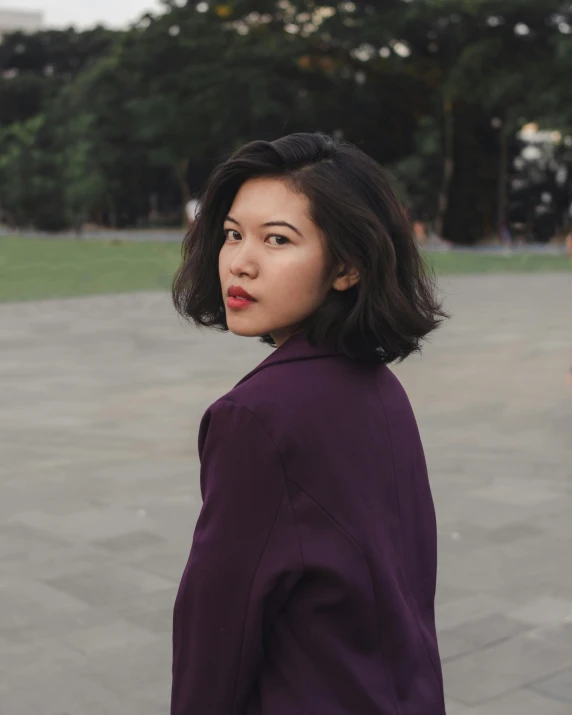 a woman standing in a park with trees in the background, an album cover, inspired by Ruth Jên, trending on unsplash, realism, a purple suit jacket, indonesia, chin-length hair, dark lipstick