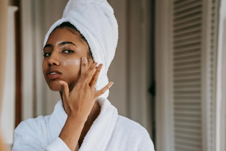 a woman standing in front of a mirror with a towel on her head, trending on pexels, manuka, early evening, hand on cheek, wearing a robe