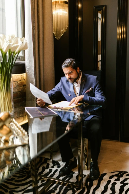 a man sitting in a chair reading a book, blue gold suit, ash thorp khyzyl saleem, on a desk, luxury lifestyle