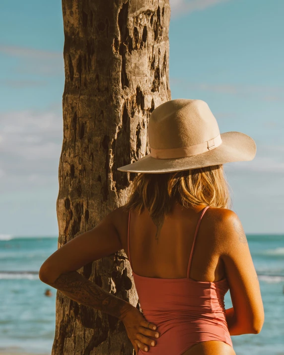 a woman standing next to a tree on a beach, pink cowboy hat, deep tan skin, tropical undertones, her back is to us