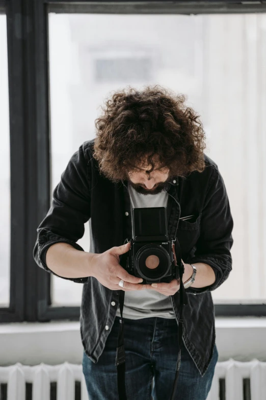 a man standing in front of a window holding a camera, curly haired, looking down on the camera, 2019 trending photo, gif