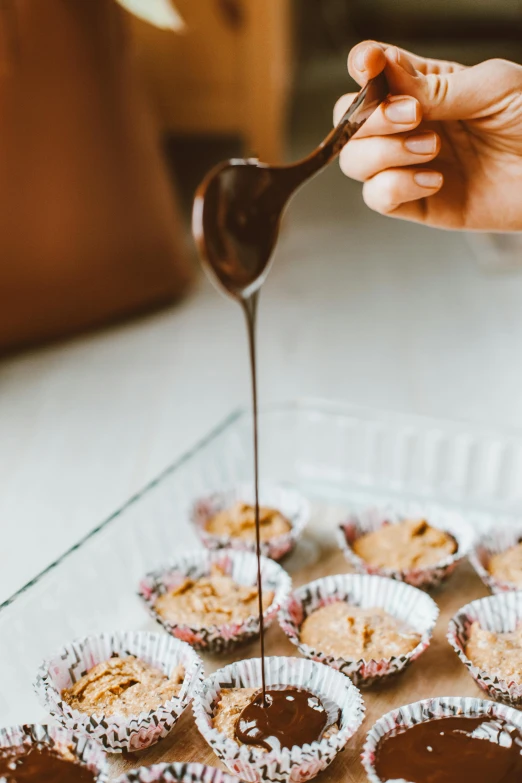 a person holding a spoon over a tray of cupcakes, syrup, fully chocolate, hammershøi, high quality product image”