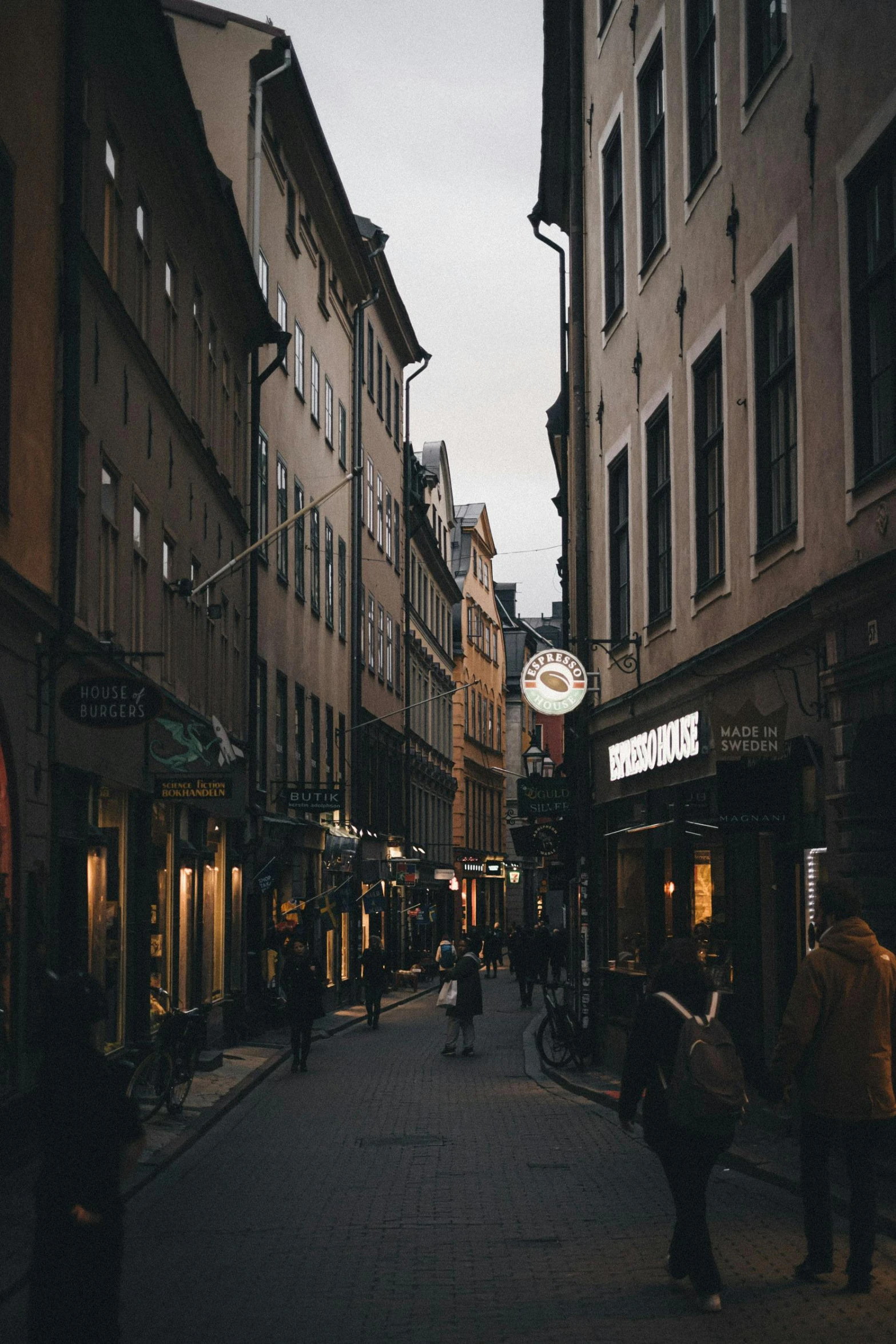 a group of people walking down a street next to tall buildings, a picture, pexels contest winner, stockholm city, warm street lights store front, colonial era street, narrow and winding cozy streets