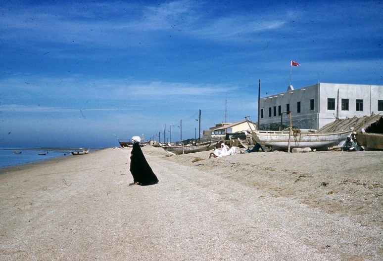 a woman in a black dress walking on a beach, a colorized photo, by Carlo Carrà, unsplash, mingei, mediterranean fisher village, islamic, mies van der rohe, slide show