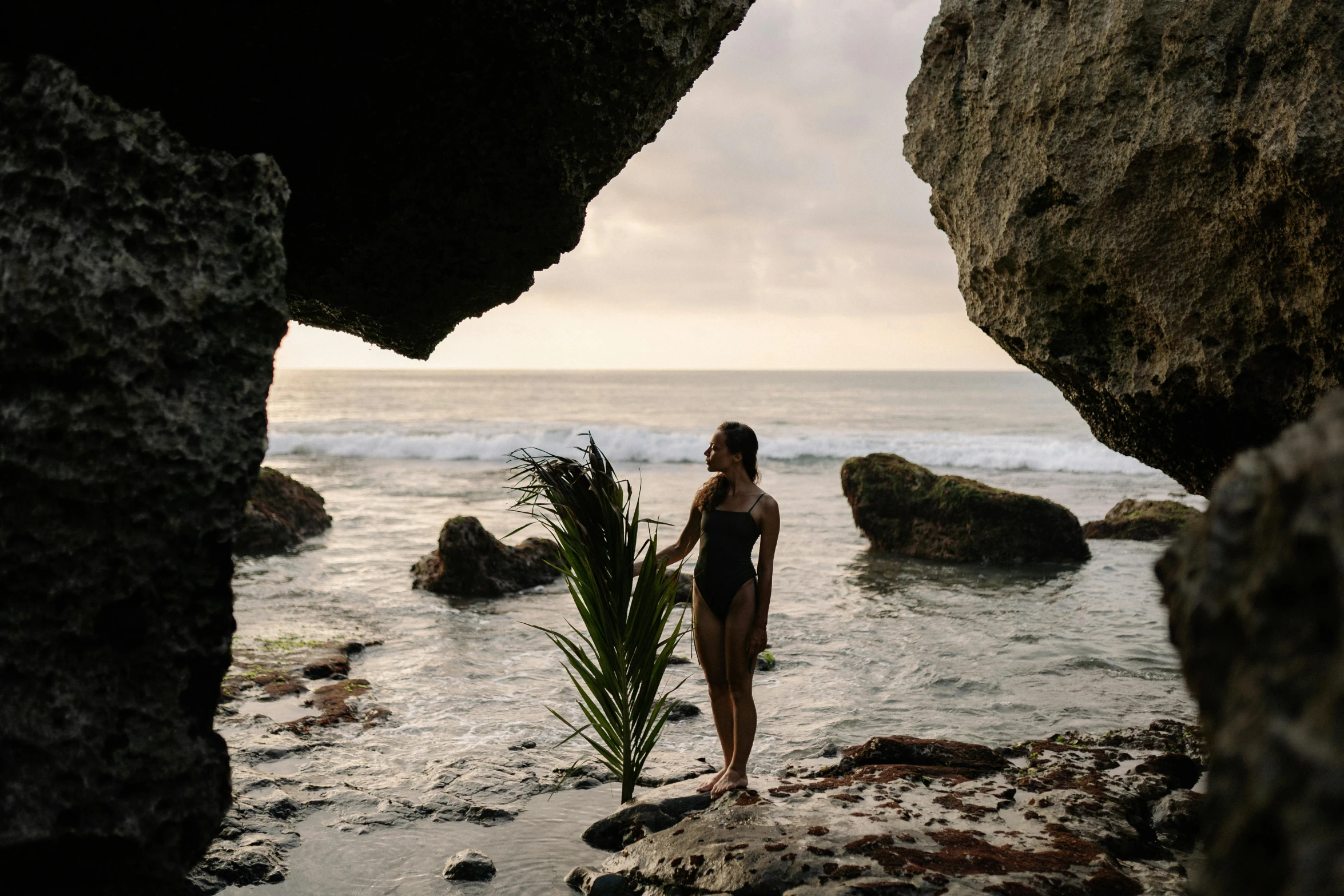 a man standing on top of a rock next to the ocean, a portrait, by Jessie Algie, unsplash contest winner, plants inside cave, woman posing, bella poarch, surf photography