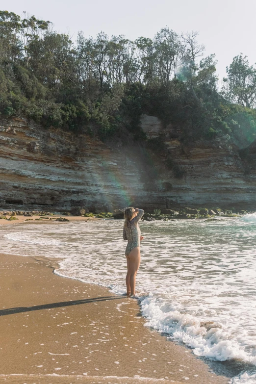 a woman standing on a beach next to the ocean, trees and cliffs, soft rainbow, bulli, sun puddle