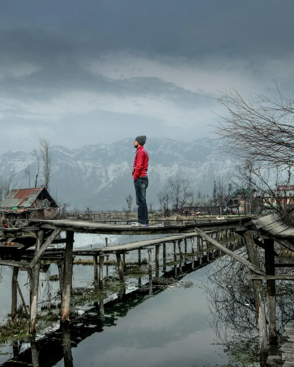 a person standing on a wooden bridge over a body of water, a matte painting, inspired by Steve McCurry, himalayas, trending photo, village in the background, lgbtq