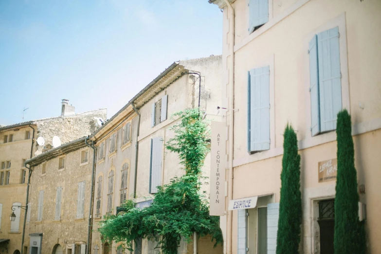 a couple of people sitting on a bench in front of a building, pexels contest winner, post-impressionism, lourmarin, pastel hues, city buildings on top of trees, summer street