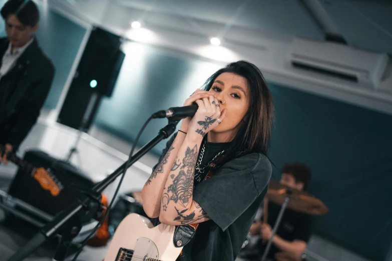 a woman singing into a microphone while holding a guitar, a tattoo, by Robbie Trevino, pexels contest winner, malaysian, looking towards camera, band playing, official store photo