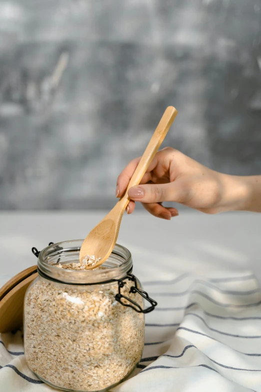 a person holding a wooden spoon over a jar of oatmeal, by Ruth Simpson, official product photo, square, light scatter, made of bamboo