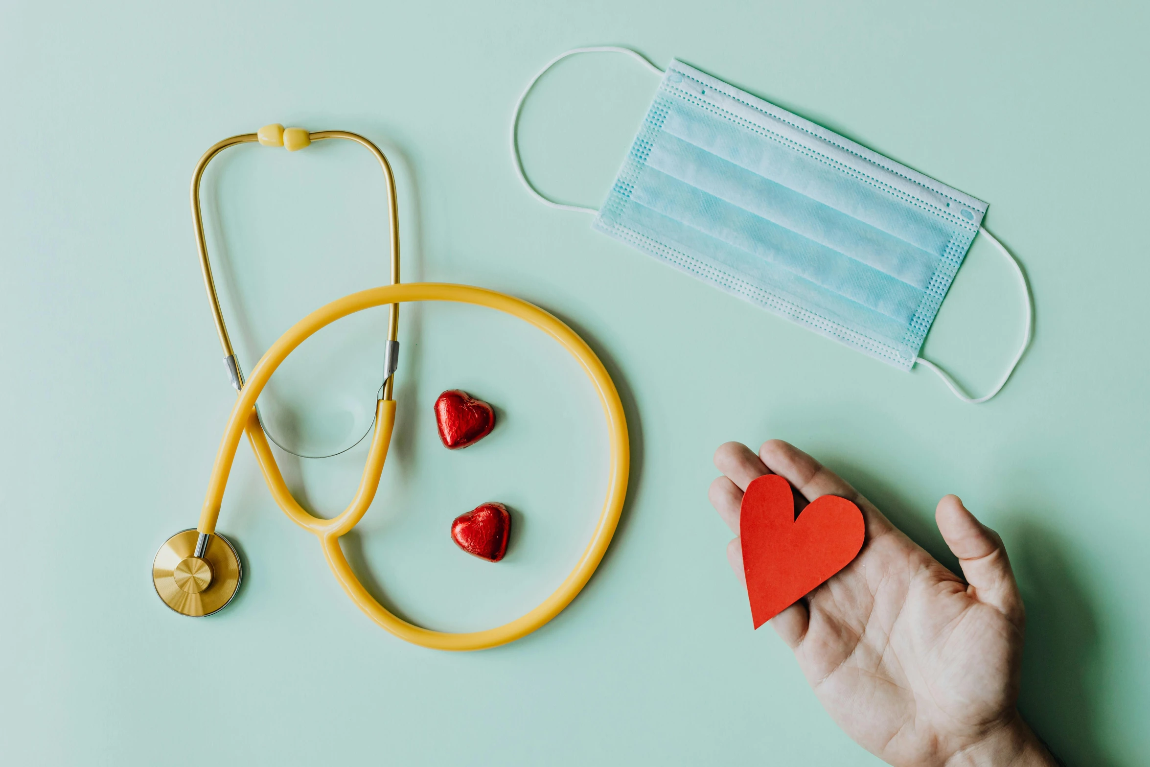 a hand holding a red heart next to a stethoscope and a stethoscope, a picture, by Julia Pishtar, trending on pexels, hurufiyya, medical mask, red and teal and yellow, 3 - piece, a green