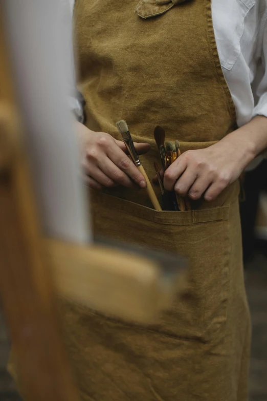 a woman that is standing in front of a easel, inspired by Kyffin Williams, pexels contest winner, blacksmith apron, holding a paintbrush in his hand, with two front pockets, ocher details