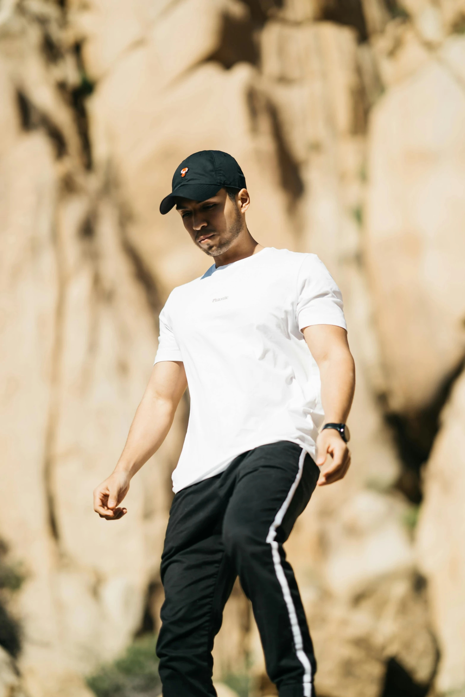 a man riding a skateboard down the side of a road, by Robbie Trevino, white baseball hat, standing on rocky ground, profile image, sport t-shirt