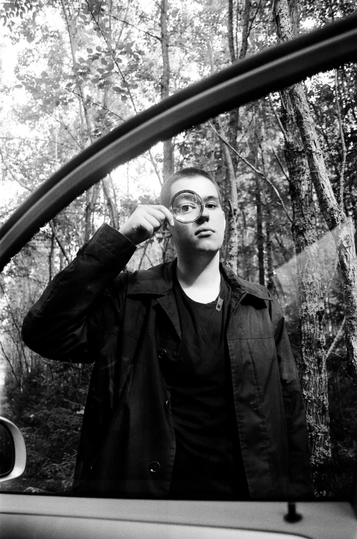 a man standing next to a car talking on a cell phone, a black and white photo, by Altichiero, brandon sanderson, standing in a forest, mac miller, eye glass