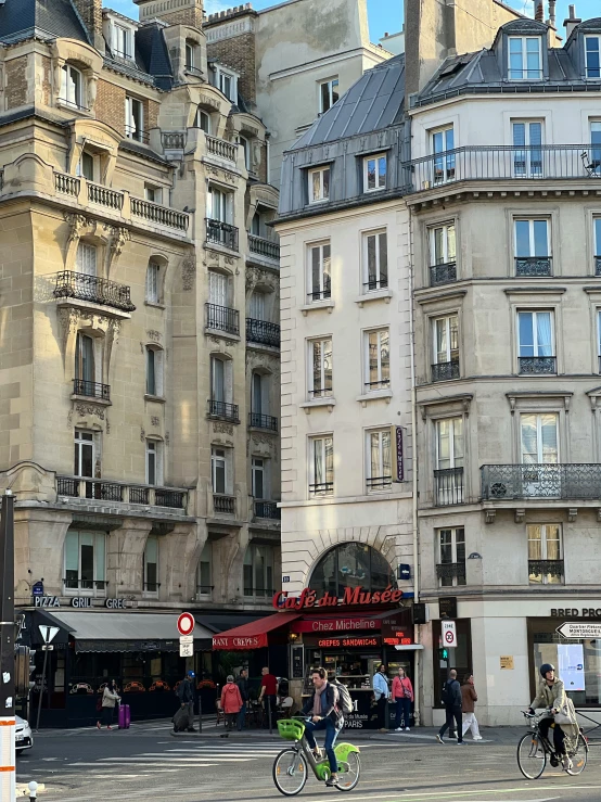 a group of people riding bikes down a street next to tall buildings, a photo, paris school, old shops, no cropping, square, illustration »