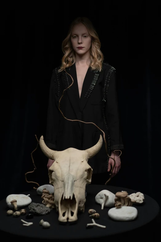 a woman standing in front of a table with skulls on it, an album cover, by Sara Saftleven, unsplash, renaissance, human dressed as a bull, black backdrop, bone and ivory, julia garner