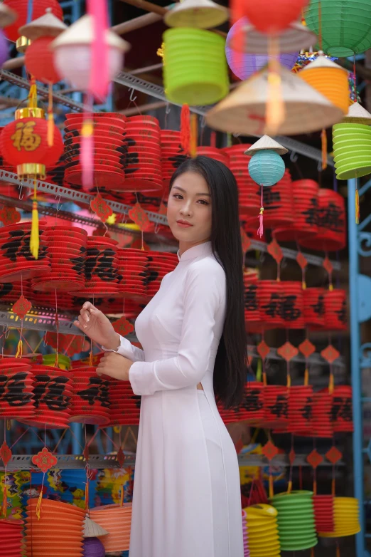 a woman standing in front of a display of lanterns, inspired by Cui Bai, pexels contest winner, young woman with long dark hair, wearing white clothes, vietnamese woman, low quality photo