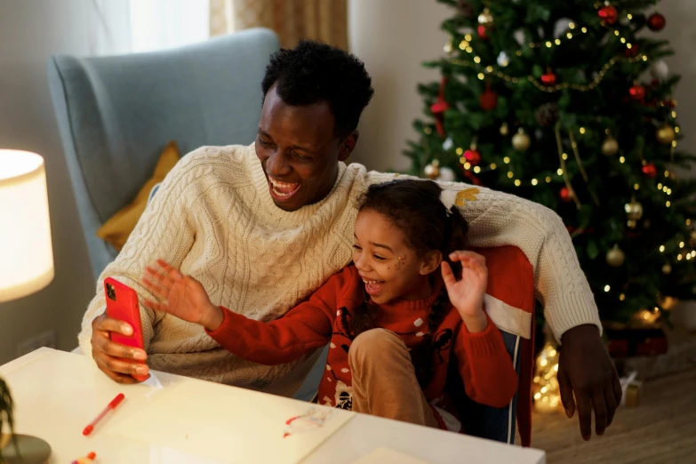 a man and a little girl sitting in front of a christmas tree, pexels contest winner, hurufiyya, looking at his phone, brown skin man with a giant grin, gif, product introduction photo