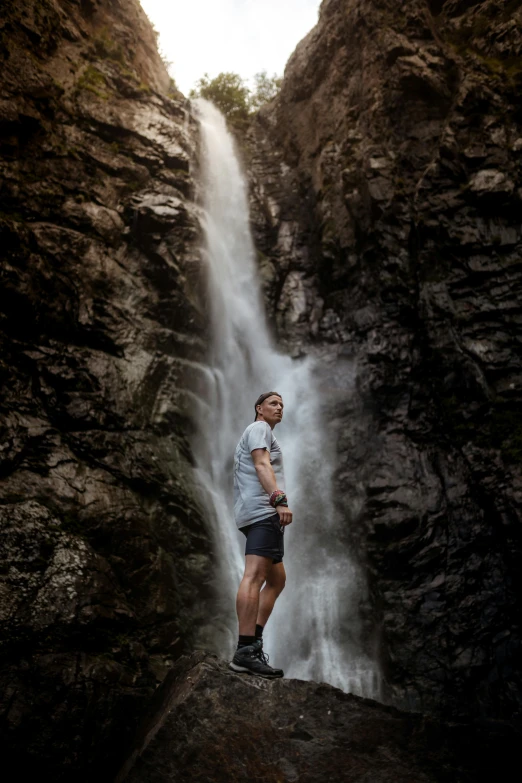 a man standing in front of a waterfall, a portrait, inspired by Michael Komarck, unsplash contest winner, white shorts and hiking boots, intense expression, 1km tall, taken in the late 2010s