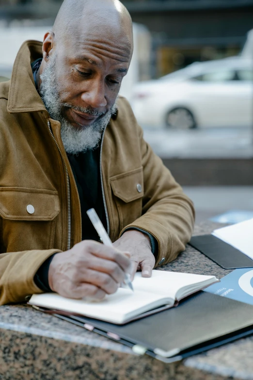 a man sitting at a table writing on a notebook, humans of new york, thumbnail, grey skinned, 2019 trending photo