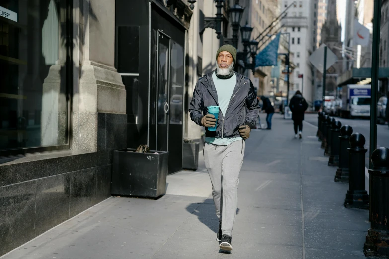 a man walking down a city street next to tall buildings, a photo, wearing teal beanie, hydration, morgan freeman, people running