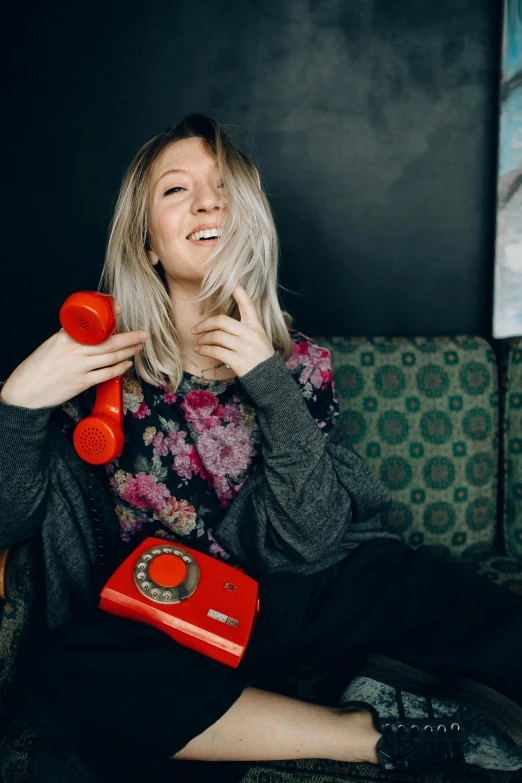 a woman sitting on a couch holding a red phone, a colorized photo, by Julia Pishtar, pexels contest winner, blonde swedish woman, playful smirk, a telephone receiver in hand, cynthwave