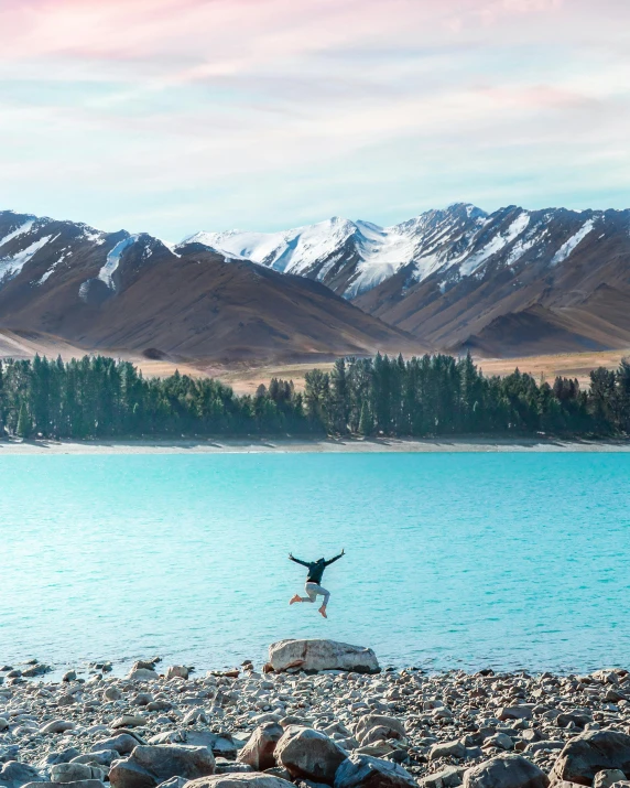 a person jumping in the air near a body of water, snowy mountains, behind that turquoise mountains, teddy fresh, new zeeland