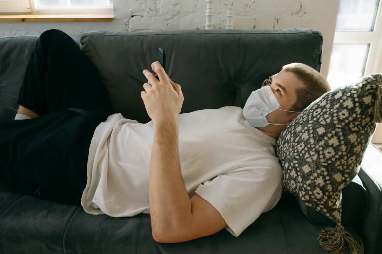 a man laying on a couch using a cell phone, trending on pexels, happening, surgical mask covering mouth, ben watts, nathan fowkes, looking across the shoulder