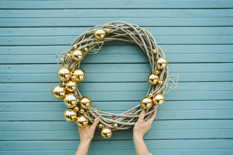 a person holding a wreath in front of a blue wall, by Julia Pishtar, pexels contest winner, golden orbs, gold cables, on grey background, wicker art