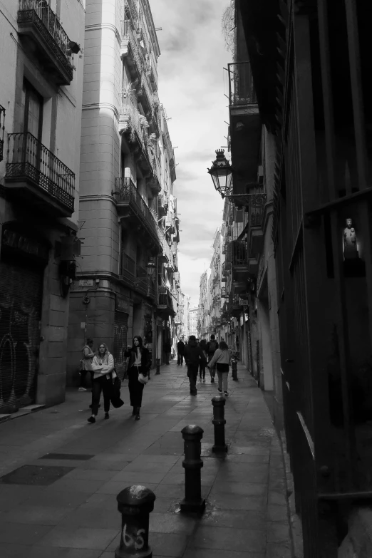 a group of people walking down a street next to tall buildings, a black and white photo, baroque, barcelona, alley, :: morning, in the evening