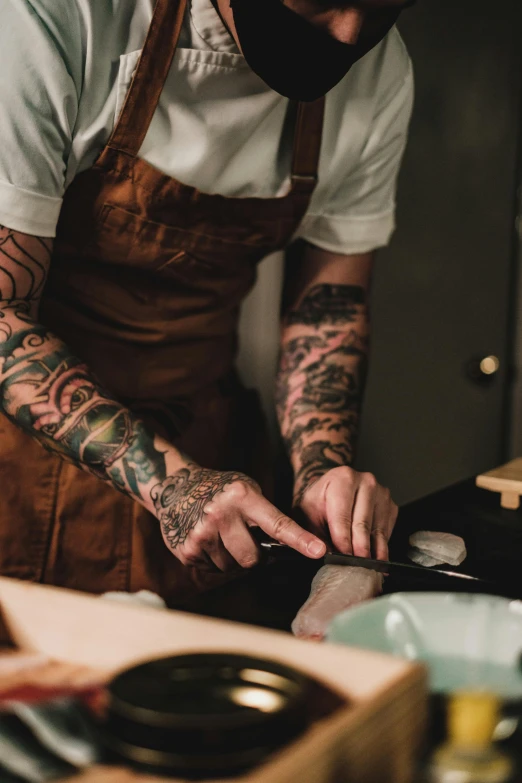 a man in an apron preparing food in a kitchen, a tattoo, trending on pexels, curved blades on each hand, aussie baristas, tattooed, knives