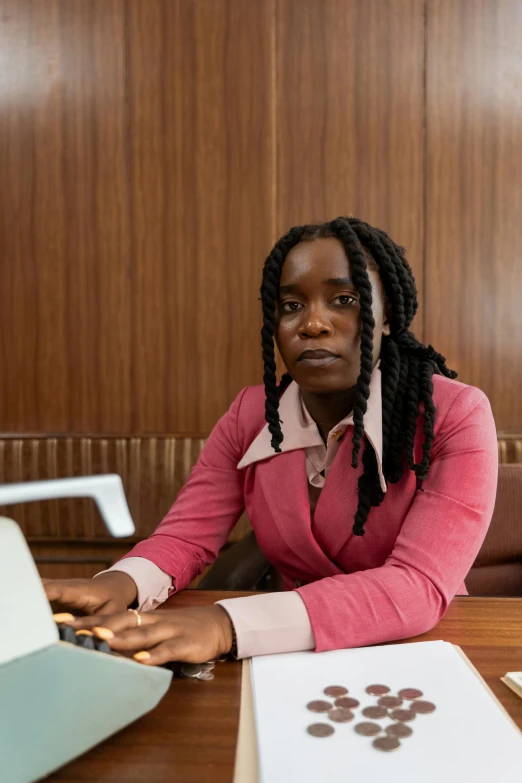 a woman sitting at a table with a laptop, by Lily Delissa Joseph, in a courtroom, adut akech, looking towards camera, plain background