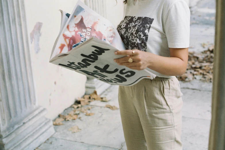 a woman standing on a sidewalk reading a newspaper, a picture, by Emma Andijewska, pexels contest winner, printed on a cream linen t-shirt, on the cover of a magazine, yes, wearing pants