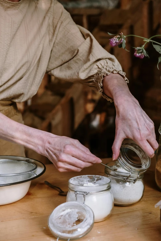 a woman putting something in a jar on a table, a still life, inspired by Elsa Beskow, trending on pexels, renaissance, female blacksmith, lined in cotton, cream, close-up photograph