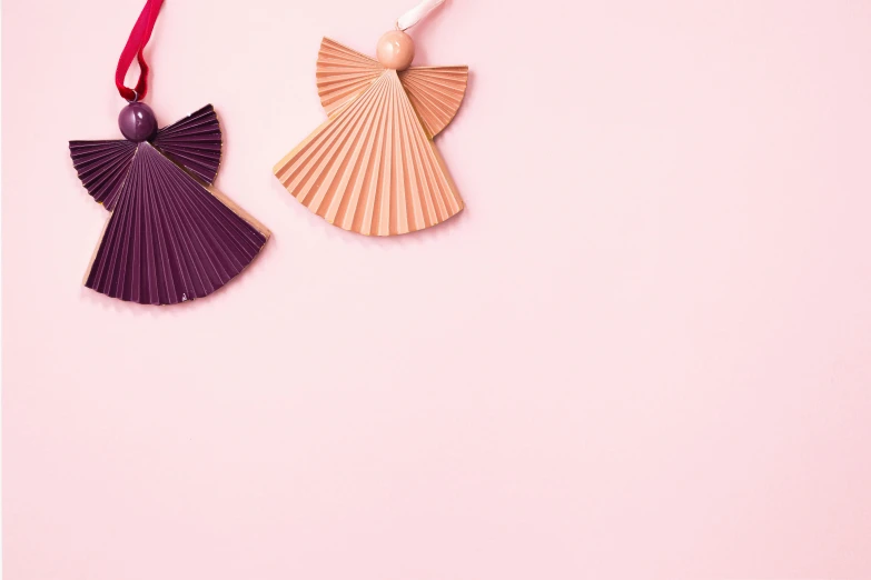 a pair of origami angel ornaments on a pink background, by Marie Angel, trending on pexels, figuration libre, hilma af klint color palette, wooden jewerly, fans hals, dressing tassels gemstones