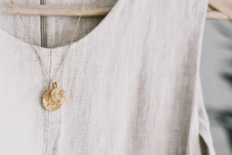 a close up of a white shirt with a gold coin on it, by Emma Andijewska, trending on pexels, minimalism, wearing several pendants, gilded. floral, linen canvas, pendant