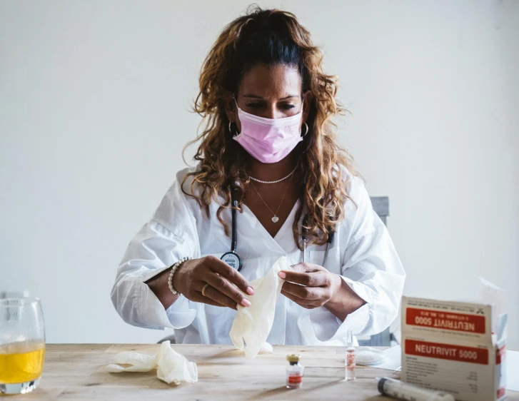 a woman wearing a face mask sitting at a table, process art, with a lab coat, first aid kit, white and pink cloth, carefully crafted