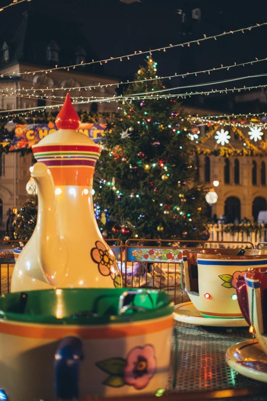 a table topped with cups and saucers next to a christmas tree, fairground rides, city square, warmly lit, zoomed out shot