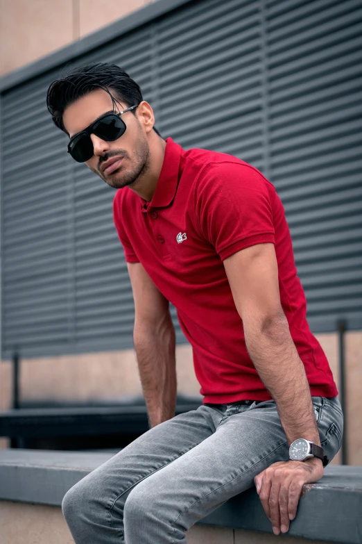 a man in a red shirt is sitting on a ledge, inspired by Nadim Karam, pexels contest winner, wearing polo shirt, model standing pose, middle eastern, wearing oakley sunglasses