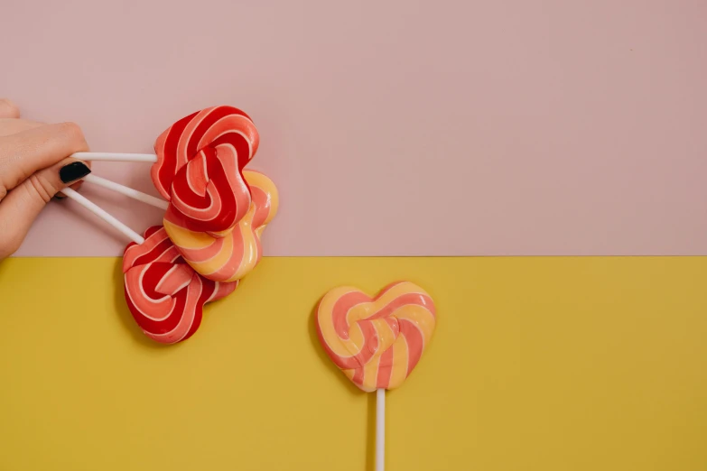 a close up of a person holding two lollipops, inspired by Ödön Márffy, trending on pexels, pop art, pink and yellow, twisted shapes, several hearts, material design