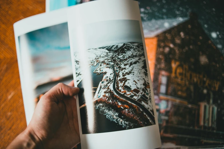 a person holding a book open on a table, a picture, pexels contest winner, art photography, “ aerial view of a mountain, risography print, lots of pictures, close up portrait photo