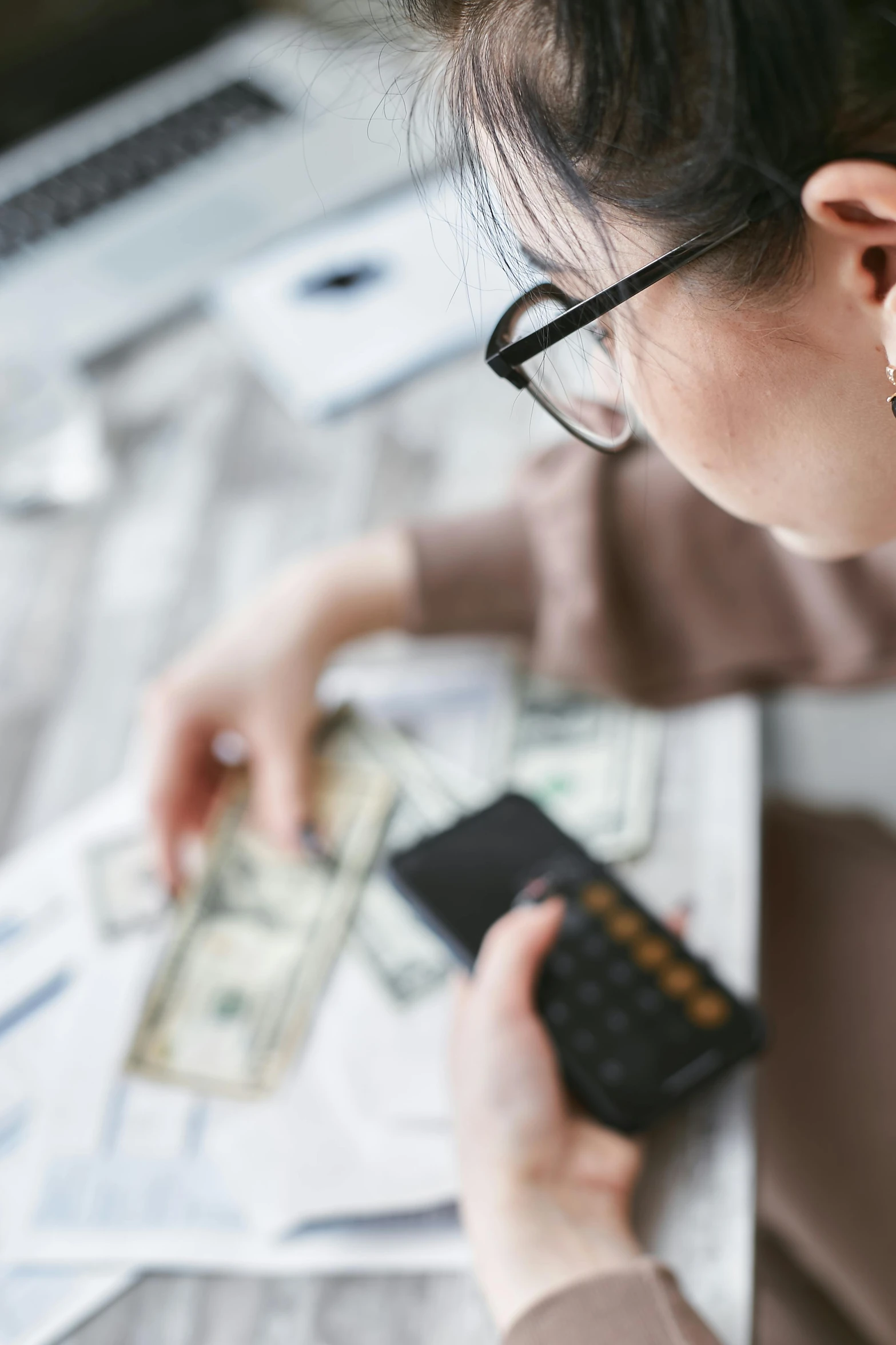 a woman sitting at a table with a calculator, pexels contest winner, money, wearing black rimmed glasses, half asian, creating a soft