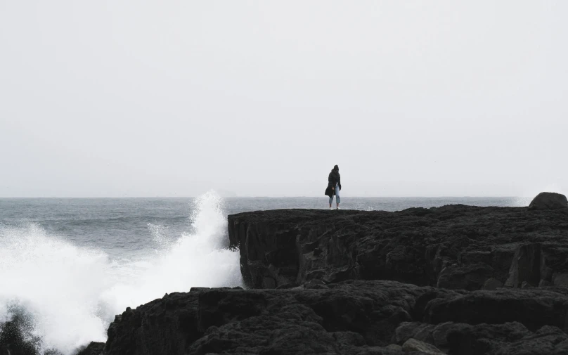 a person standing on top of a cliff next to the ocean, pexels contest winner, postminimalism, waves crashing at rocks, a lonely woman, grey, the seventh seal