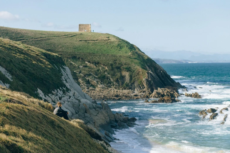 a couple of people sitting on top of a hill next to the ocean, by Lee Loughridge, unsplash contest winner, renaissance, old ruins tower, bulli, dezeen, manuka