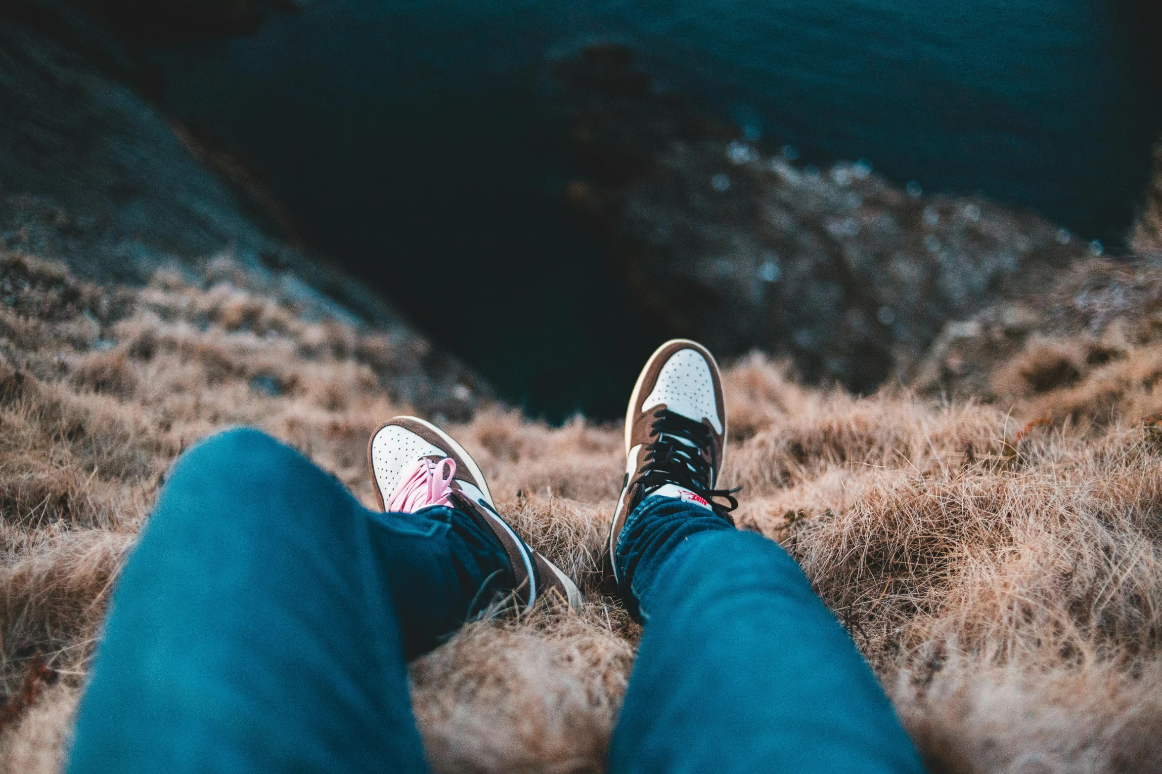 a person sitting on top of a hill next to a body of water, trending on pexels, tight blue jeans and cool shoes, lo fi colors, looking up at camera, high cliff