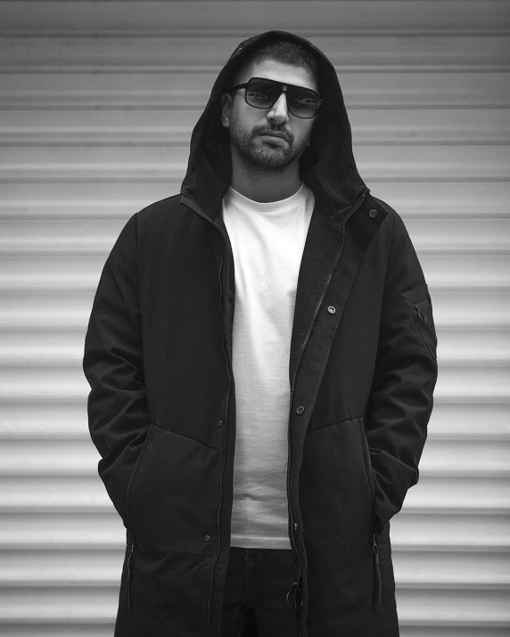 a man standing in front of a garage door, a black and white photo, lyco art, dressed black hoodie, wearing shades, long black jacket, artem chebokha
