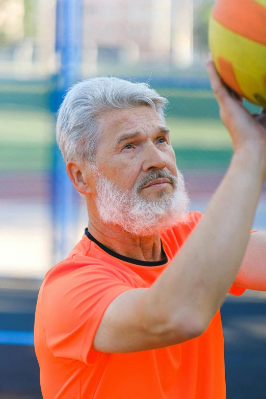 a man in an orange shirt holding a yellow and orange ball, gray hair and beard, square, 5 5 yo, volleyball