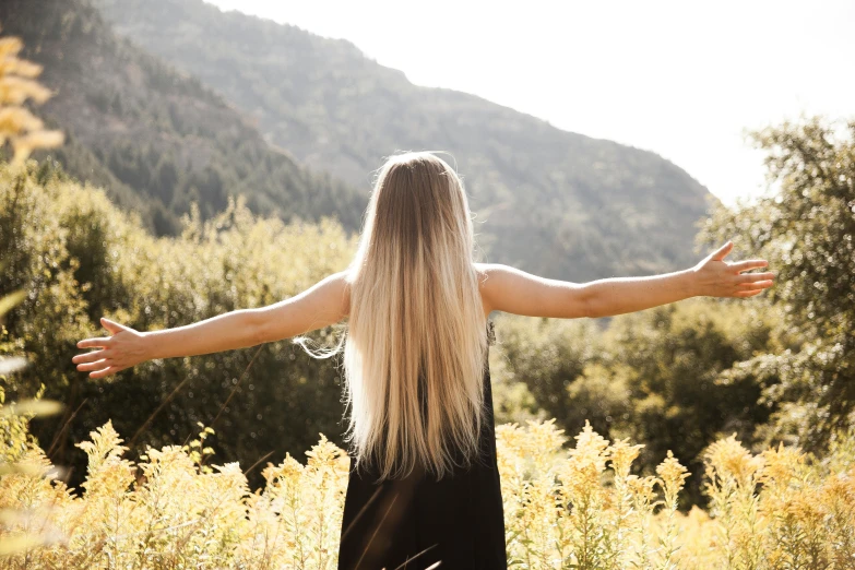 a woman standing in a field with her arms outstretched, trending on unsplash, long luxurious light blond hair, overlooking a valley, styled hair, back - lit