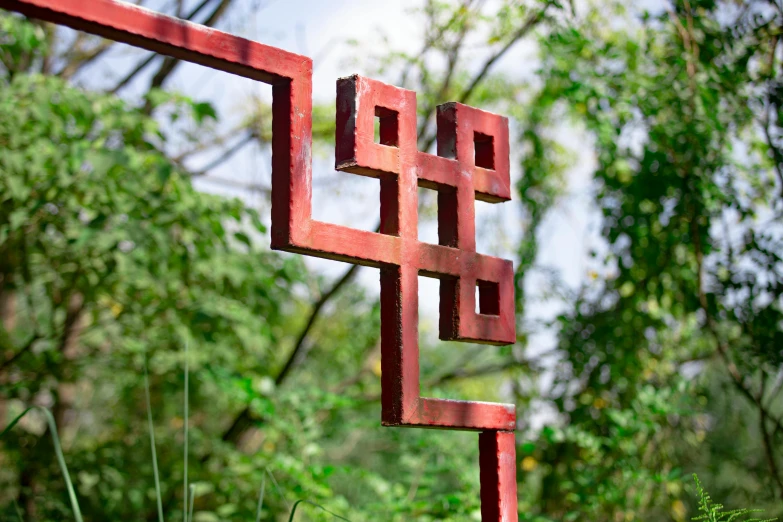 a close up of a metal cross with trees in the background, an abstract sculpture, inspired by Torii Kiyomoto, unsplash, kinetic art, red iron oxide, serpentine maze, squared border, devanagari script